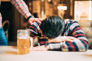 Young man in casual clothes is sleeping near the mug of beer on a table in pub, another man is waking him up. Get drunk man.