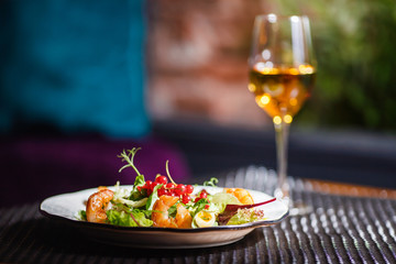 A plate with a beautiful appetizing salad of fresh vegetables, shrimps, quail eggs and lingonberries is on the table with a drink in a wine glass.