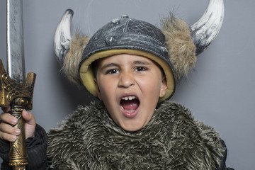 viking brunette boy in fur shirt in preschool age standing indoors, wearing costume hat with horns, hands up, making a scary face and sword