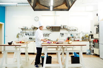 Young chef of restaurant standing by table and cutting and other vegetables for stew