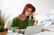 © shurkin_son - Picture of beautiful casually dressed European student girl with red hair having bored expression while doing research project at home, sitting at workplace with laptop, phone and textbooks on desk