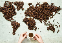 Hands Hold A Cup Of Coffee Against The Background Of The World Map From Coffee Beans