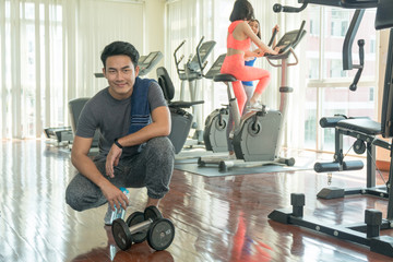 Muscular young man with dumbbell and towel resting after workout with blurred young woman slim body on the fitness machines background in gym. Exercise and sport concept