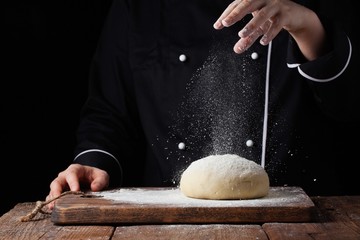 Chef hands pouring flour powder on raw dough using sieve on a black background, Cooking process