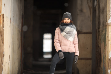 Wall Mural - A young cute white girl in a hat warm scarf and gloves is standing in the corridor of a ruined house in winter. Destroyed building.