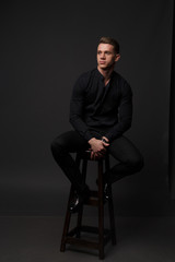 Wall Mural - A white man sits on a dark chair in a black shirt and black mills, on a gray background