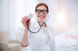 © zinkevych - Doctors tool. Appealing skillful experienced female doctor staying on blurred background while grinning and rising stethoscope