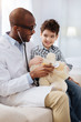 © Viacheslav Yakobchuk - Next patient. Gay optimistic male doctor sitting with boy while examining plush bear and laughing