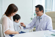 © DragonImages - Middle-aged doctor looking at his cute little patient with wide smile while examining him with stethoscope, interior of modern office on background