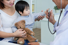 Curious Asian Boy Sitting On Laps Of His Pretty Mother And Examining Stethoscope With Interest While Having Consultation At Pediatrician Office