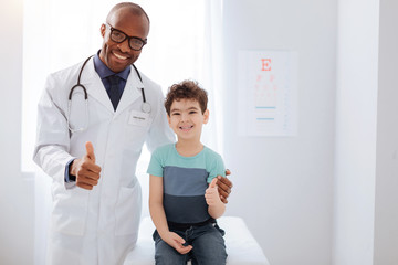 Wall Mural - Excellent health. Cheerful happy male doctor and boy smiling while looking at camera and showing thumbs