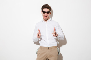 Sticker - Perky man in black sunglasses pointing index fingers on camera with lots of money dollar bills sticking out from pockets, isolated over white background with shadow