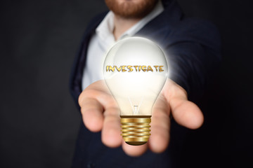 A businessman is holding a light bulb with the inscription:INVESTIGATE