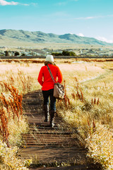 woman in red coat walking along wooden path in high grassy field