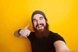 Fototapeta  - Cheerful smiling bearded hipster man showing thumbs up and making selfie over yellow background