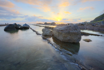 Wall Mural - Sunrise scenery of a rocky coast in northern Taiwan, with beautiful reflections of golden sunlight on peaceful sea water under dramatic dawning sky (long exposure) ~ Scenic view of a beach in twilight