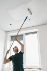 Wall Mural - a man with a roller in his hand on a long stick paints the ceiling in gray at the window. Repair in a bright room.