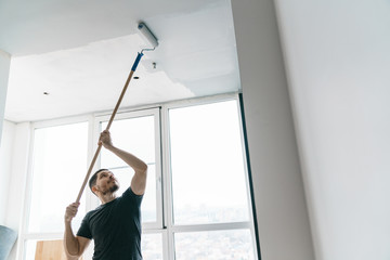 Wall Mural - The man paints the walls and the ceiling in gray on his balcony in the backlight. Painting and repair in the room.