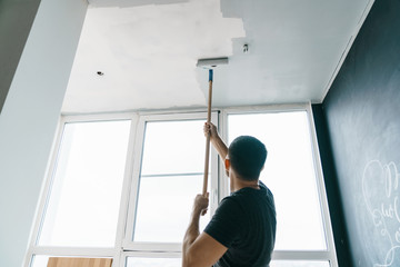 Wall Mural - the man paints the walls and the ceiling in gray color, standing with his back to the camera. Focus on the roller. Painting and repair of the room.