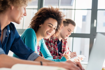 Cheerful young woman writing an assignment while sitting at desk between two classmates during class at college or university