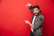 Photo of agitated man wih beard in casual clothing pointing fingers on copyspace text or product with surprise and smile, isolated over red background