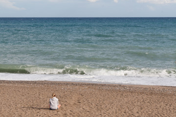 Warm clear sunny summer day. A girl in a gray sweatshirt and jeans shorts sits with her back to the camera on the sad shore and looks at the sea