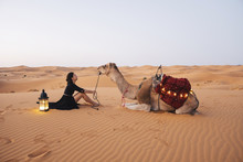 Side View Of Woman With Camel Sitting On Sand At Sahara Desert Against Clear Sky