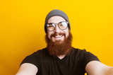 Fototapeta  - Cheerful bearded hipster an with eyelgasses taking selfie in studio