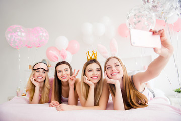 Wall Mural - Cheerful, joyful, charming, pretty, funny, dreamy, cute models lying on bed shooting self portrait on front camera, holding using smart phone, showing two fingers, peace symbol, enjoying theme party