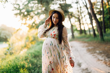 pregnant girl in a long embroidered beige dress and a straw hat stands on a forest road against a sunset background