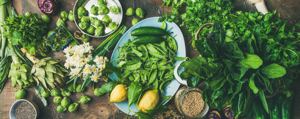 Spring healthy vegan food cooking ingredients. Flat-lay of vegetables, fruit, seeds, sprouts, flowers, greens over wooden background, top view, wide composition. Diet, clean eating food concept