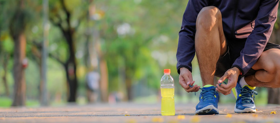 Young athlete man tying running shoes in the park outdoor, male runner ready for jogging on the road outside, asian Fitness walking and exercise on footpath in morning. wellness and sport concepts