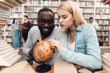 Wall Mural - Ethnic african american guy and white girl surrounded by books in library. Students are using globe.