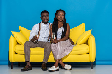 Wall Mural - beautiful young african american couple holding wine glasses and smiling at camera while sitting on yellow sofa