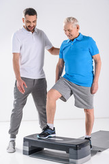 rehabilitation therapist helping senior man exercising on stepper isolated on grey