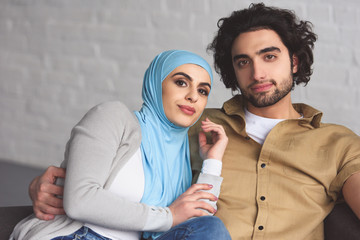 Canvas Print - happy muslim couple sitting on sofa in living room and looking at camera