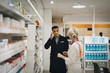 © Maskot - Male customer explaining headache to female pharmacist standing by rack at store