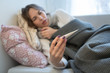 © DimaBerlin - Sick young Caucasian woman lying on sofa,  wrapped in plaid blanket and scarf, having flu symptoms, holding and looking at thermometer, focus on thermometer/Cold, virus, sickness concept/Flu season