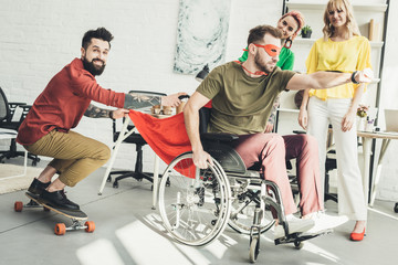 disabled businessman in superhero costume with colleague on skateboard and businesswomen behind in office