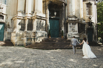 Wall Mural - Elegant wedding couple is walking in old ancient town to the church. Slim bride in satin dress with train and groom in grey checkered suit. Photo on cobblestone road near the medieval cathedral.