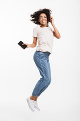 Wall Mural - Full length image of happy african american woman with brown curly hair wearing jeans and t-shirt levitating with smartphone in hand, isolated over white background