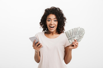 Wall Mural - Happy winner american woman with afro hairstyle and big smile holding money prize dollar cash and silver smartphone, isolated over white background