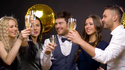 Wall Mural - celebration, luxury and holidays concept - happy friends with golden balloons clinking champagne glasses at party over black background