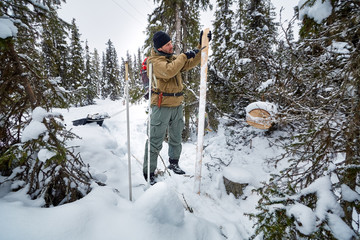 A young man cleans wooden skis covered with snow with knife in winter  tundra forest.