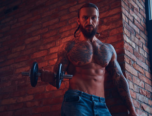 A handsome tattoed shirtless male with a stylish haircut and beard, doing exercise with a dumbbell, standing against a brick wall in a room with a loft interior.