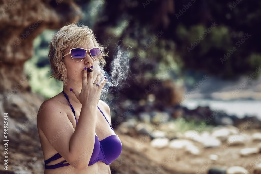 Blonde woman smoking a cigar. Blonde woman in bikini smoking a cigar at the  beach. Stock Photo