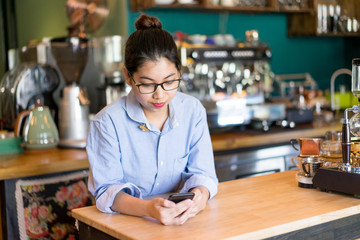 Serious coffee shop owner reading message on phone while waiting for clients. Busy young Asian female entrepreneur using smartphone at workplace. Modern small business concept