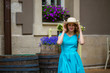 © Елена Кравчук - Young woman smiling looking camera, in blue dress, holding hat, outdoor, beautiful city, purple flowers