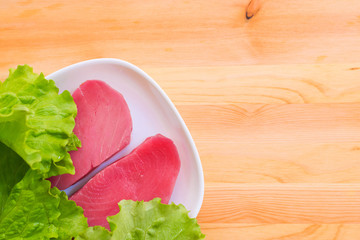 Two pieces of raw fresh tuna fillet on a plate with large leaves of a green salad on a wooden table. Delicious sea food easy. Greek cuisine