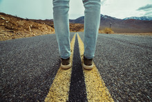 Men`s Feet Standing On Aslphalt Desert Road With Yellow Marking Lines. Man Wearing Sneakers And Jeans.